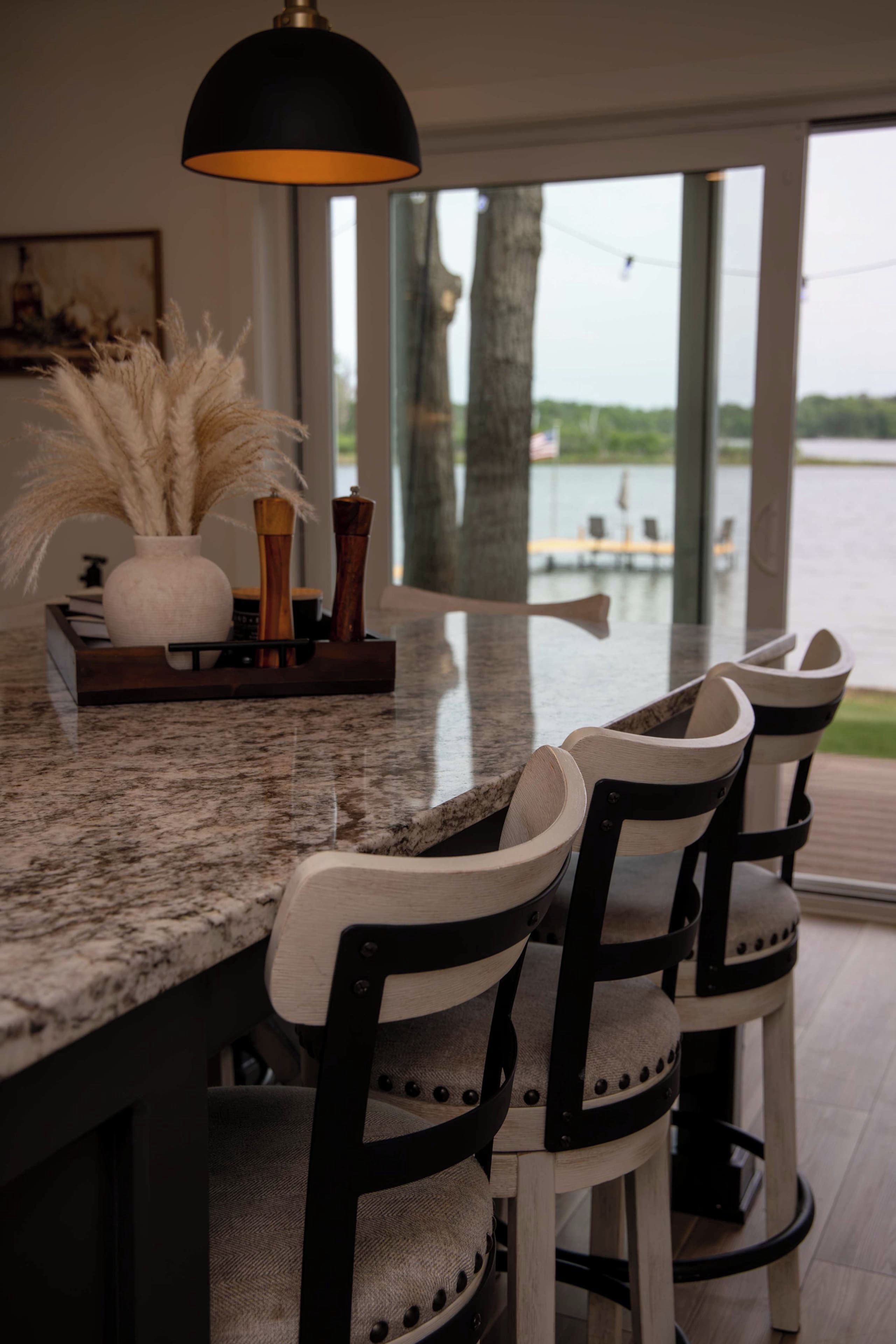 A kitchen with a granite countertop, a hanging light fixture, a vase with dried plants, and a view of a body of water through large windows.