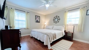 Un dormitorio con cama, ventilador de techo, escritorio, silla, cubo de basura y ventana con cortinas.