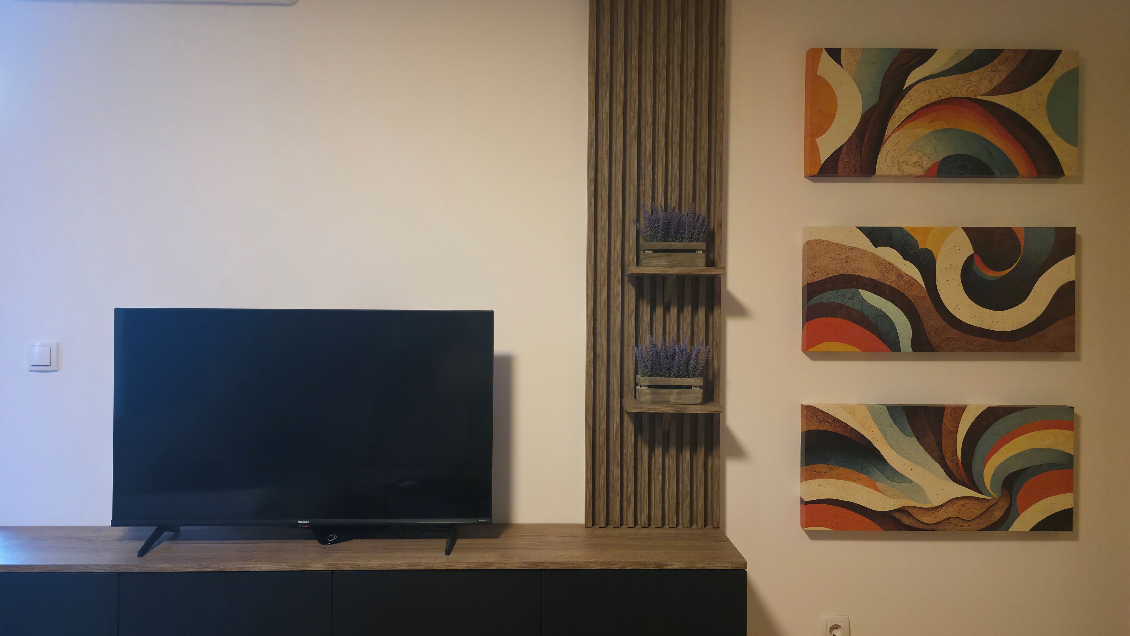 Un televisor de pantalla plana sobre un soporte de madera, flanqueado por dos estantes fijos a la pared con objetos decorativos y tres pinturas abstractas.