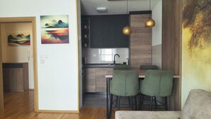 Een moderne keuken met een donker aanrecht, een spoelbak en twee groene barkrukken. Er hangen twee schilderijen aan de muur, één toont een berglandschap en de ander een kleurrijk abstract ontwerp.