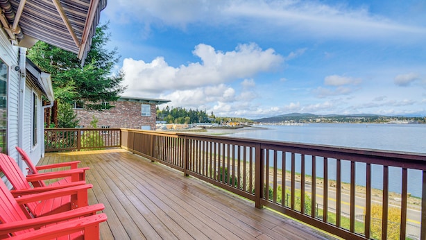 A wooden deck with red chairs, a stone building, and a view of a lake and distant mountains. - Walk to Beaches: Waterfront Home in Port Orchard! (Port Orchard)