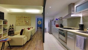 Une cuisine moderne équipée d’appareils électroménagers en acier inoxydable, un coin repas avec un canapé et une table, et un salon avec une télévision et des étagères à livres.