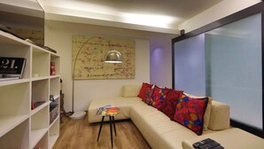 Un salon moderne avec un canapé beige, des coussins colorés, une petite table d’appoint, une lampe sur pied et une bibliothèque.