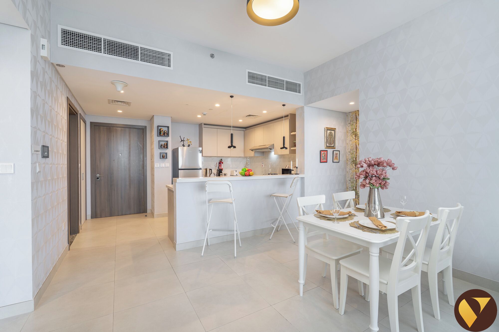 A modern kitchen with white cabinetry, a central island, and pendant lighting. There is a dining area with a white table and chairs, and a vase with flowers on the table.