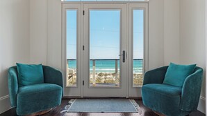 Una habitación con un gran ventanal corredera que da a un balcón con vista al océano. Hay dos sillones color turquesa y una alfombra azul en el suelo.