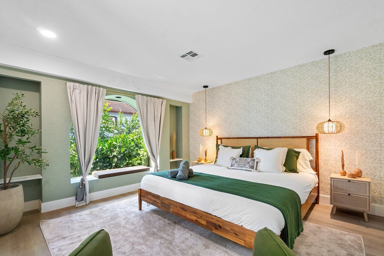 A bedroom with a large bed, a bedside table, a nightstand, a window with curtains, a potted plant, and a view of greenery outside.