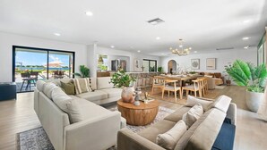 A spacious living area with a large sofa, a coffee table, and a dining table with chairs. There is a large window providing a view of the outdoors. - The Garden of Eden | Beautiful Villa w/ Pool & Game Room (West Palm Beach)