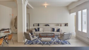 A spacious living room with a white sofa, a glass coffee table, and a patterned rug. There is a dining area with a table and chairs, and a large window providing natural light.