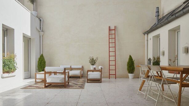 A spacious indoor area with a high ceiling, a wooden table with chairs, a red ladder, and several potted plants.
