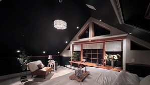 A bedroom with a large bed, a chandelier, a window with plants, and a small table with a lamp.