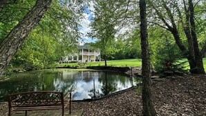 A serene park with a pond, a bench, and a house in the background.