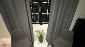 Een kamer met twee grijze stoelen, een witte bijzettafel met een plant erop, en uitzicht door een raam met metalen spijlen.