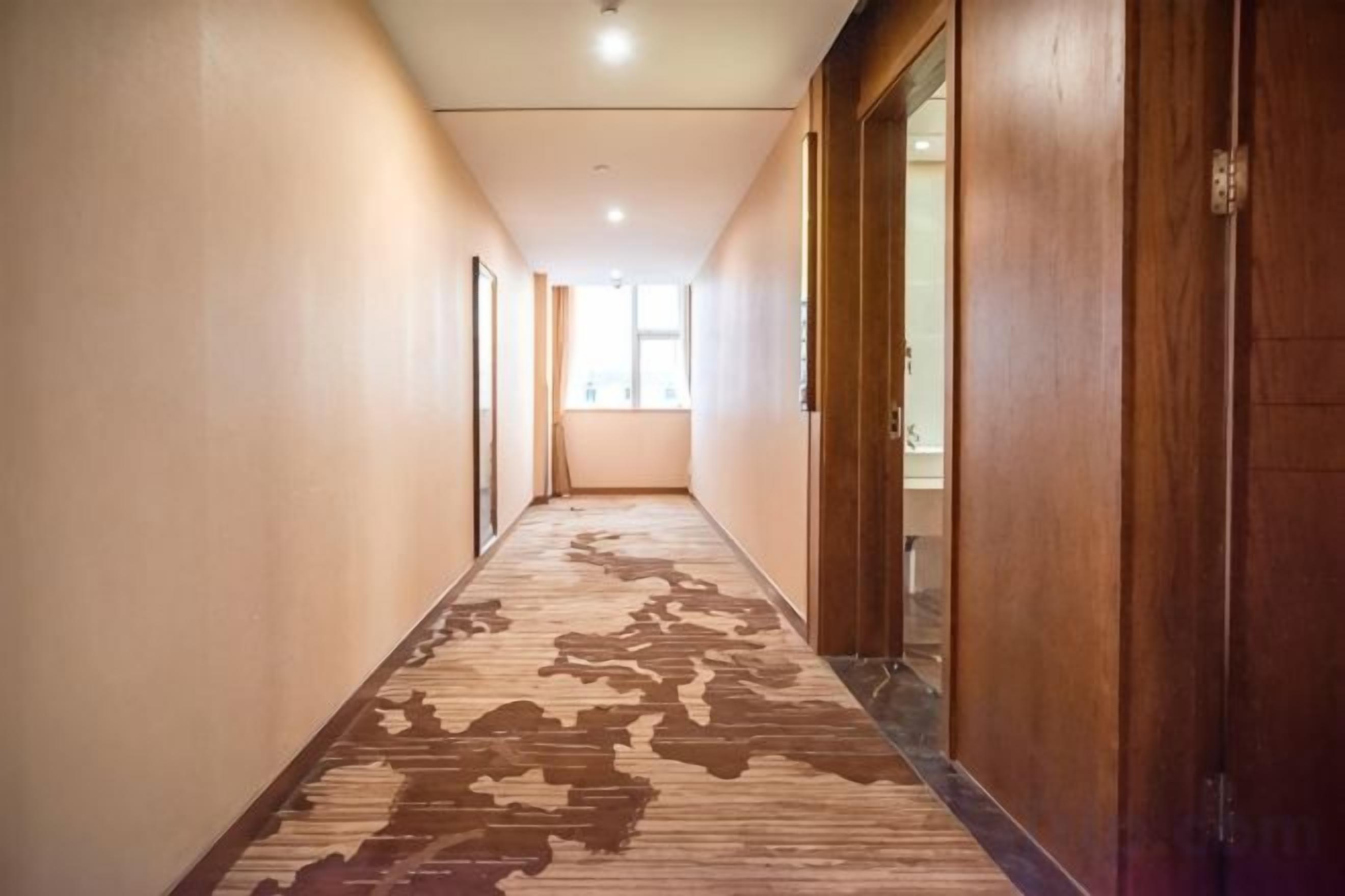 A hotel corridor with carpeted flooring, wooden doors, and a window at the end.