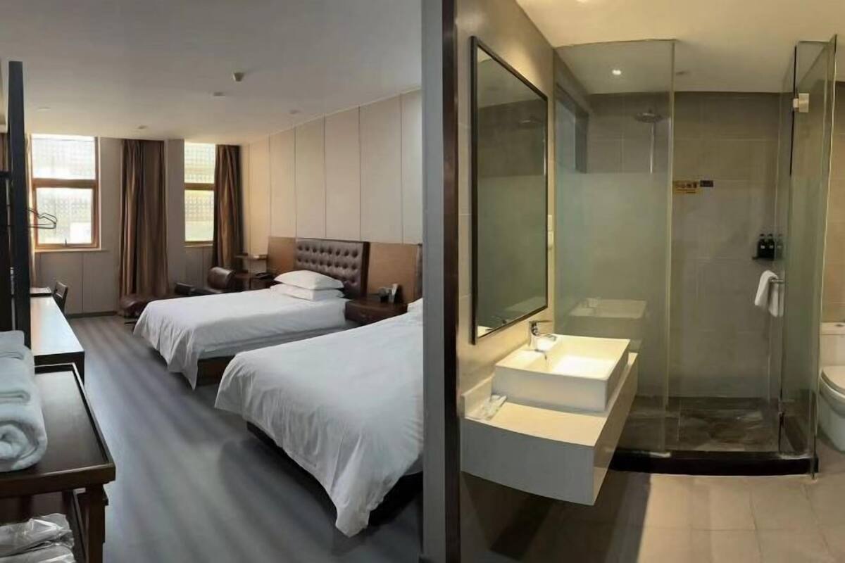 A hotel room with two beds, a desk, a large mirror, and a glass-enclosed shower area.