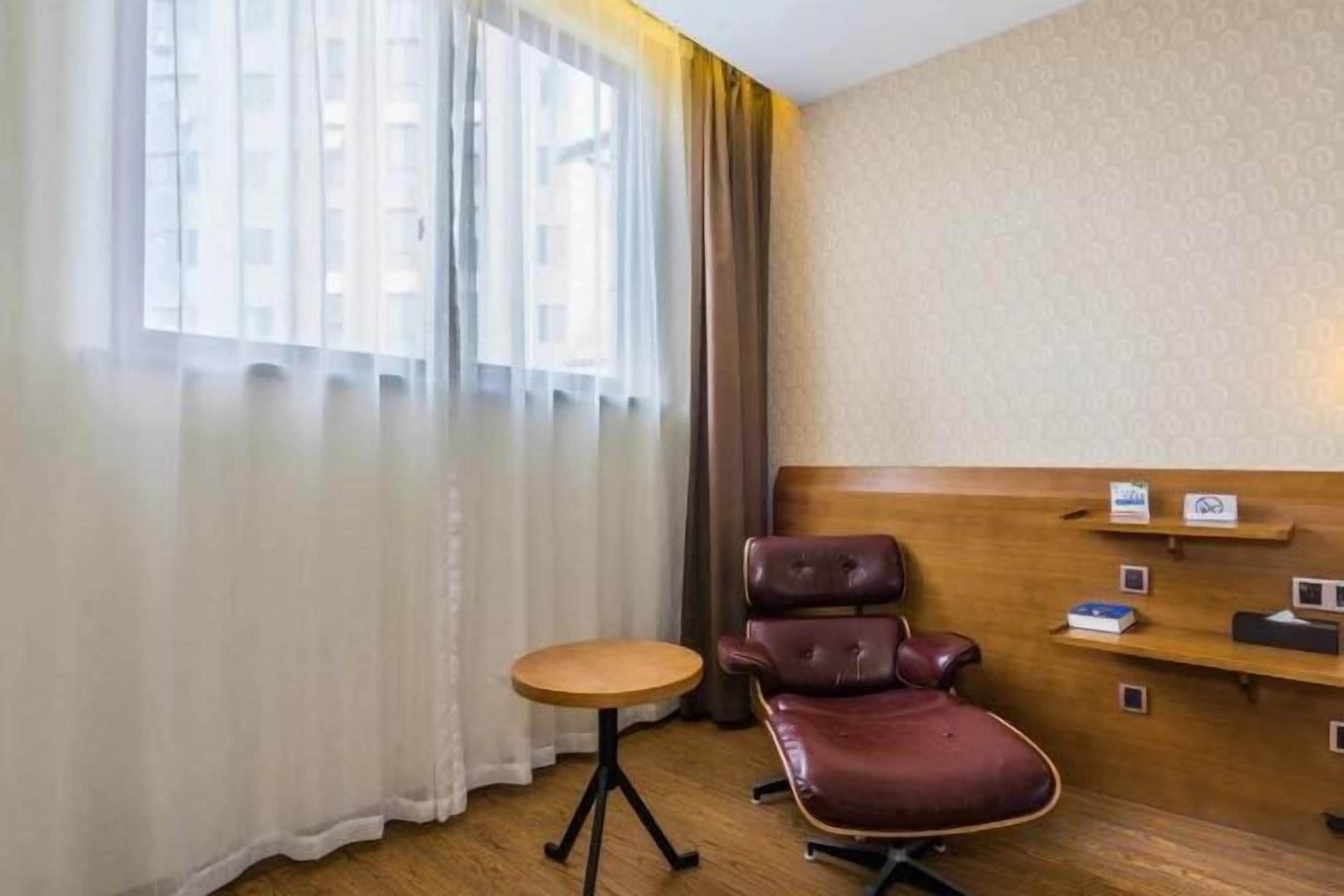 Hotellihuone, jossa on puinen sängynpääty, pieni pyöreä pöytä, nahkainen tuoli ja ikkuna, jossa on ohuet verhot.