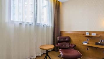 Hotellihuone, jossa on puinen sängynpääty, pieni pyöreä pöytä, nahkainen tuoli ja ikkuna, jossa on ohuet verhot.