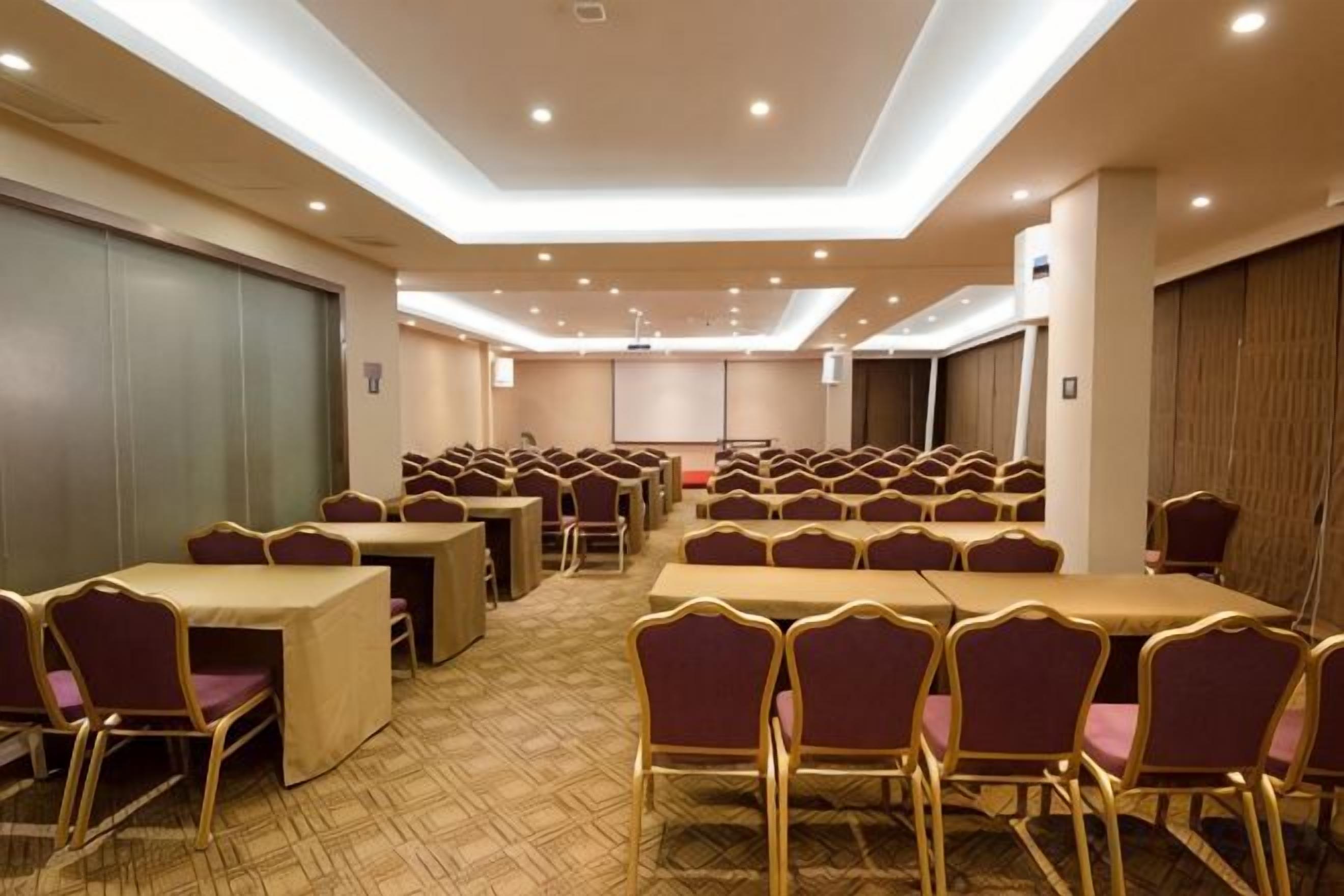 A conference room with rows of tables and chairs, a projector screen, and a carpeted floor.