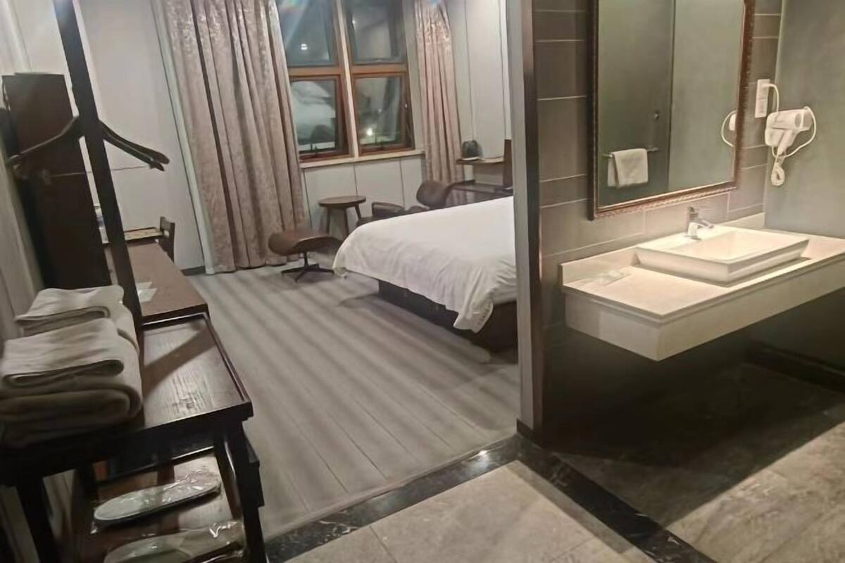 A hotel room with a bed, a desk with towels, a bathroom with a sink and mirror, and a window with curtains.