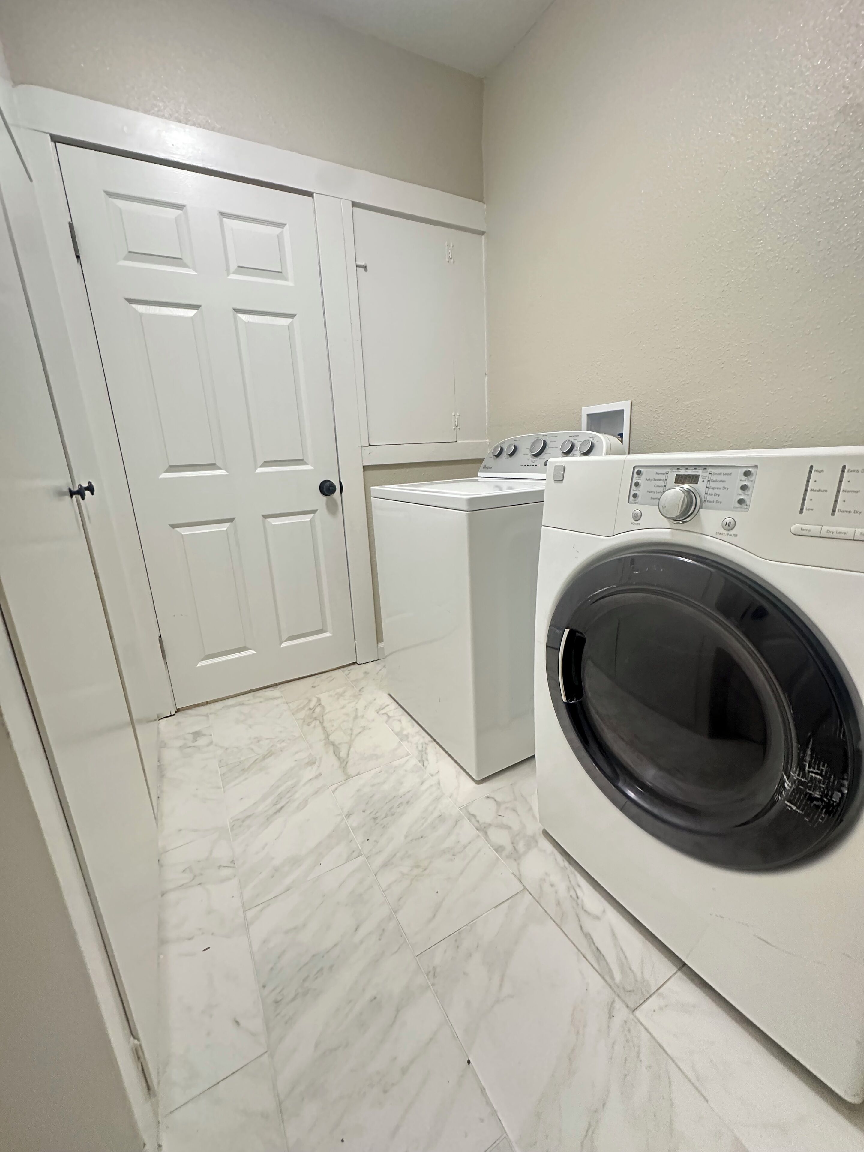 A laundry room with a front-loading washer and dryer, a white door, and a tiled floor.