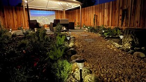 A covered outdoor seating area with a view of a landscape, surrounded by a wooden fence and a garden with rocks and plants.