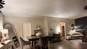 A modern living room with a dining area, featuring a wooden floor, a dining table with chairs, a sofa, and a large wall clock.