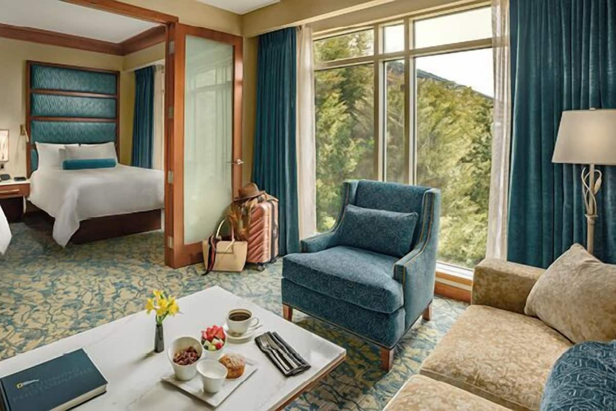 Habitación de hotel con cama, sofá, sillón, mesa de centro con alimentos para el desayuno y vistas a zonas verdes.