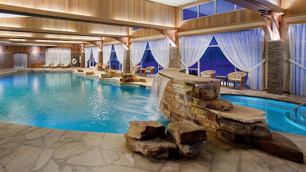 An indoor swimming pool with a waterfall feature, wooden deck seating, and stone walls. - The Lodge at Turning Stone Resort Casino (Verona)