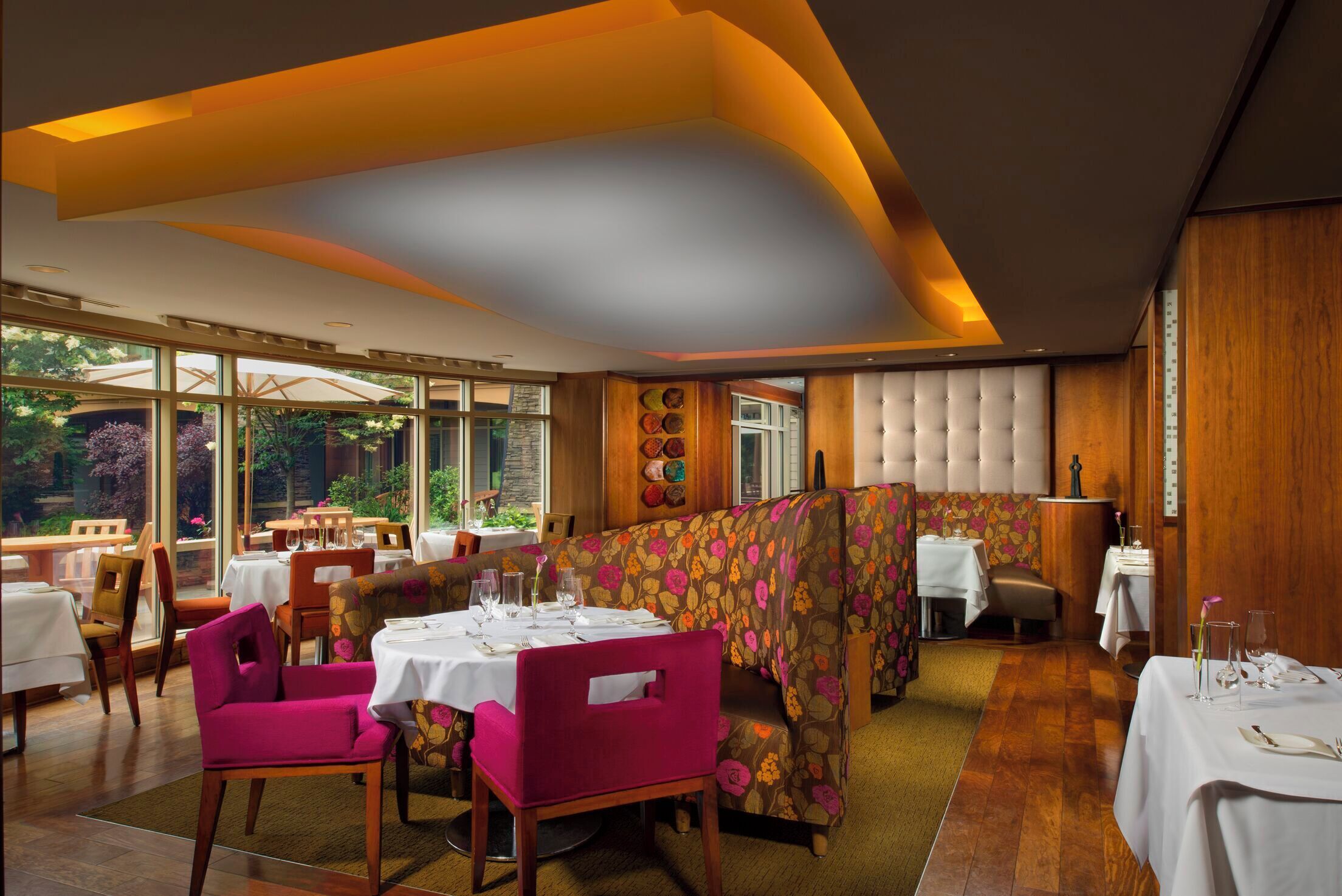 Un restaurant avec un plafond haut, de grandes fenêtres et un paravent décoré de motifs floraux. Les tables sont dressées avec des nappes blanches et des chaises rouges.