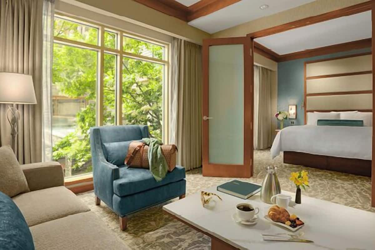 Habitación de hotel con un sillón azul, una mesita de café blanca con provisiones para el desayuno, una cama con colcha blanca y vistas a la vegetación a través de amplias ventanas.