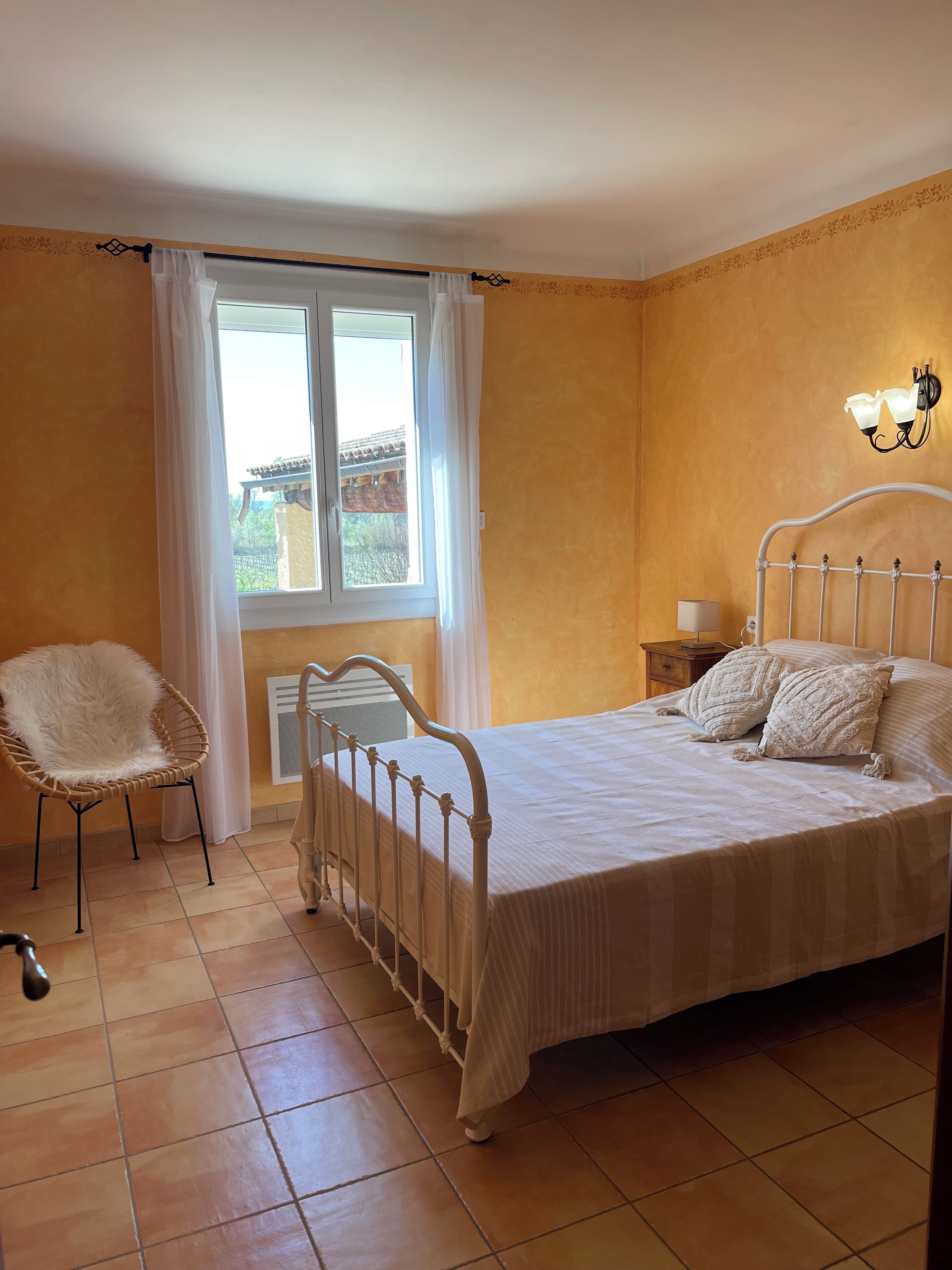Un dormitorio con una cama, una silla, una mesita de noche, una lámpara y una ventana con cortinas.