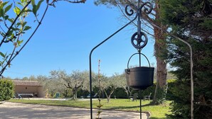 Ein Steinbrunnen mit einem Metallkübel, der an einer Spiralenkonstruktion hängt, umgeben von Grünpflanzen und einem Gebäude im Hintergrund.