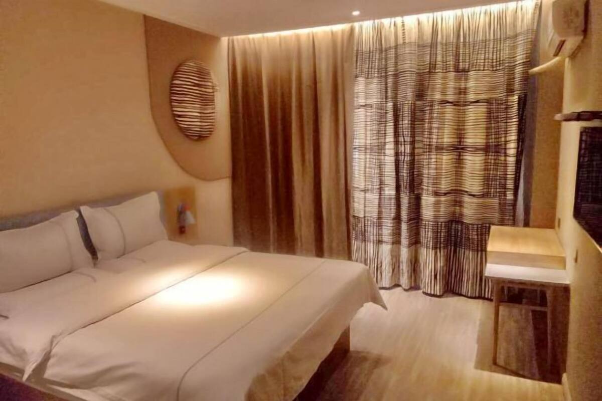 A hotel room with a bed, a bedside table, a wall-mounted TV, and a window with curtains.