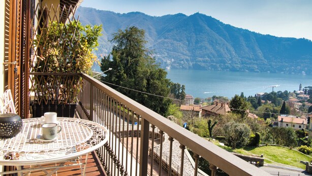 A balcony with a table, potted plants, and a view of a lake and mountains.