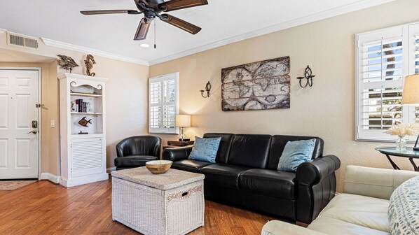 A living room with a black leather sofa, a white wicker ottoman, a globe artwork on the wall, and a ceiling fan.