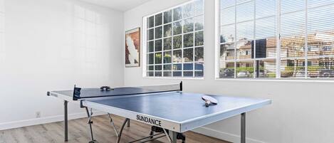 A ping pong table in a room with large windows, wooden flooring, and a white wall.
