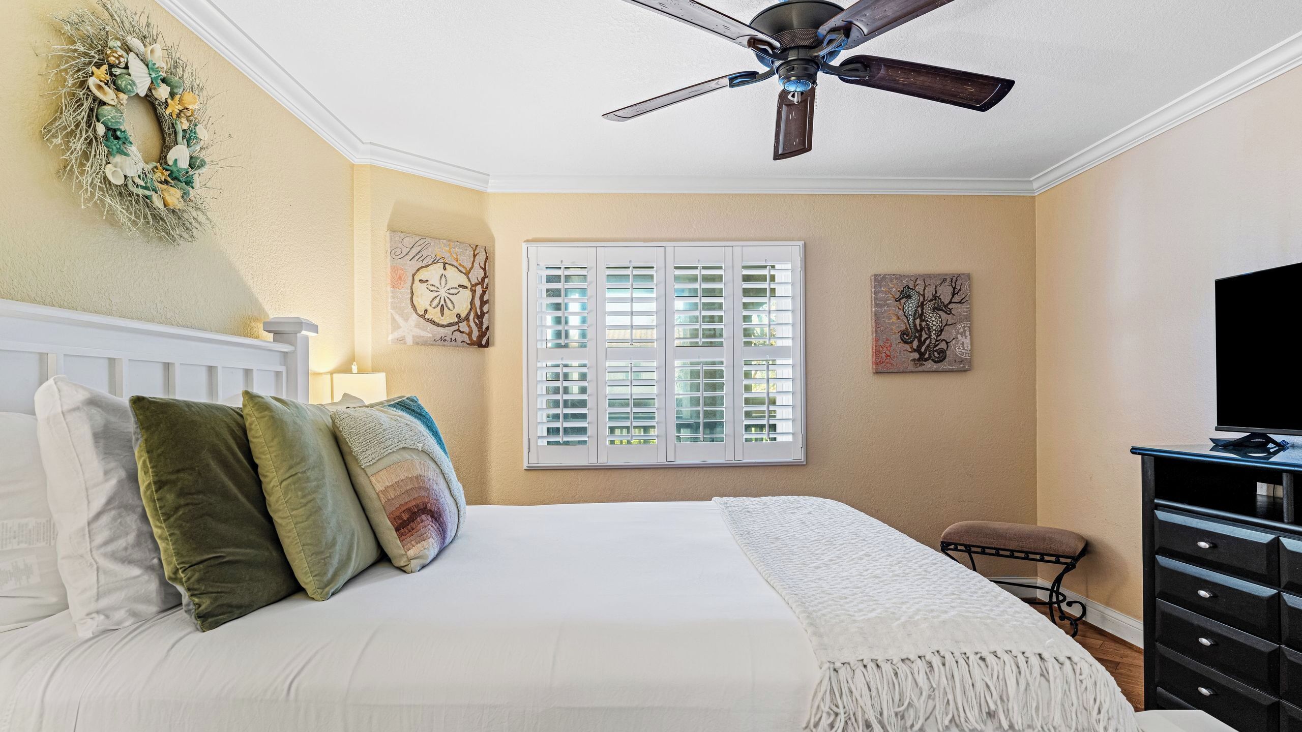 A bedroom with a bed, pillows, a ceiling fan, a window with shutters, a dresser, and artwork on the walls.