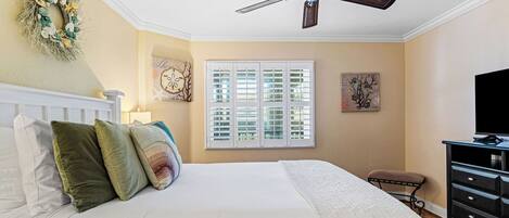 A bedroom with a bed, pillows, a ceiling fan, a window with shutters, a dresser, and artwork on the walls.
