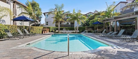 A rectangular swimming pool with a tiled deck, surrounded by lounge chairs and umbrellas, with palm trees and a building in the background.