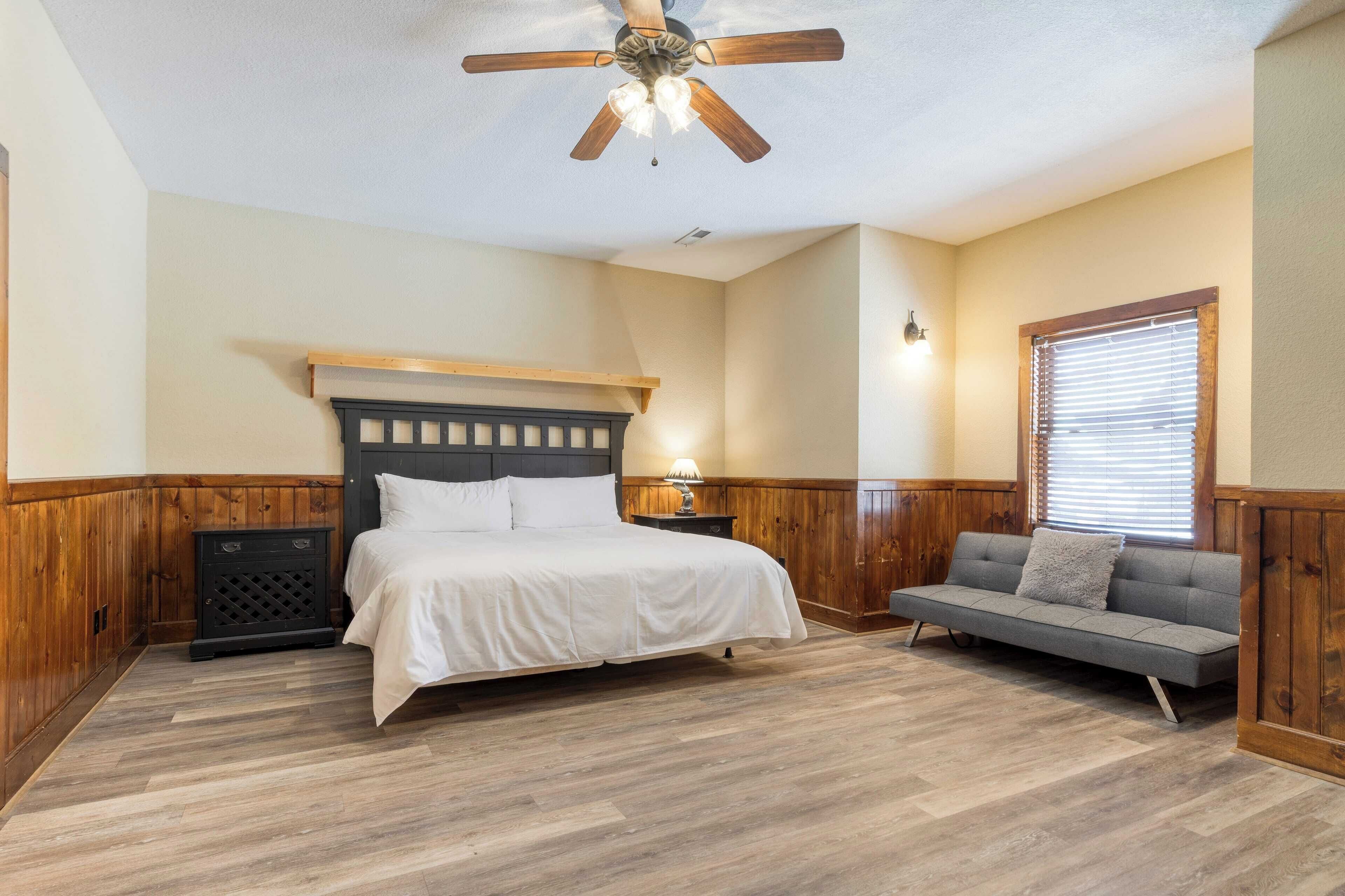 A bedroom with a bed, a nightstand, a sofa, a ceiling fan, and a window with blinds.