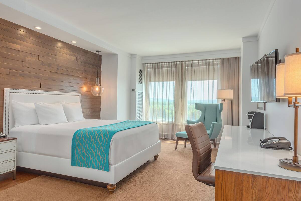 A hotel room with a wooden accent wall, a bed with white bedding, a desk with a lamp, and a chair.