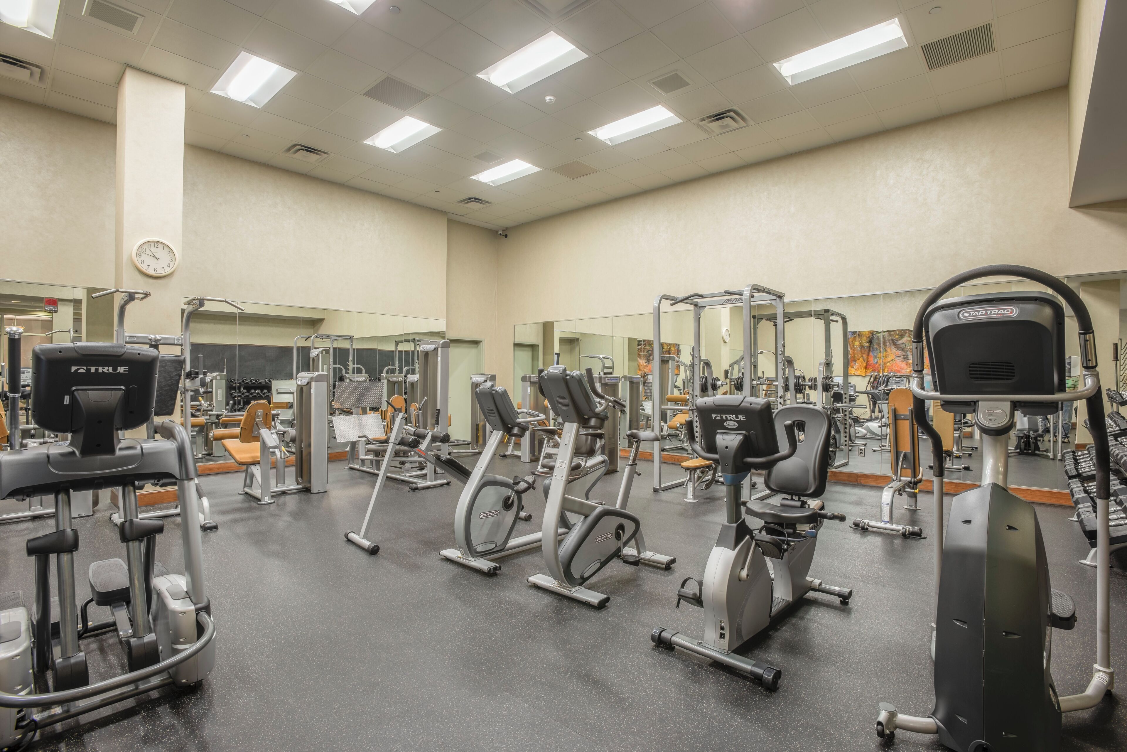 Un gimnasio con diferentes máquinas de ejercicio, espejos y un reloj en la pared.