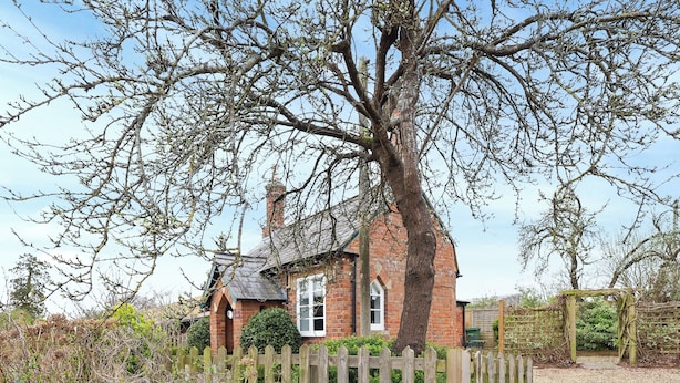 Une maison traditionnelle en briques, avec un grand arbre devant, entourée d’une clôture en bois et de diverses plantes.