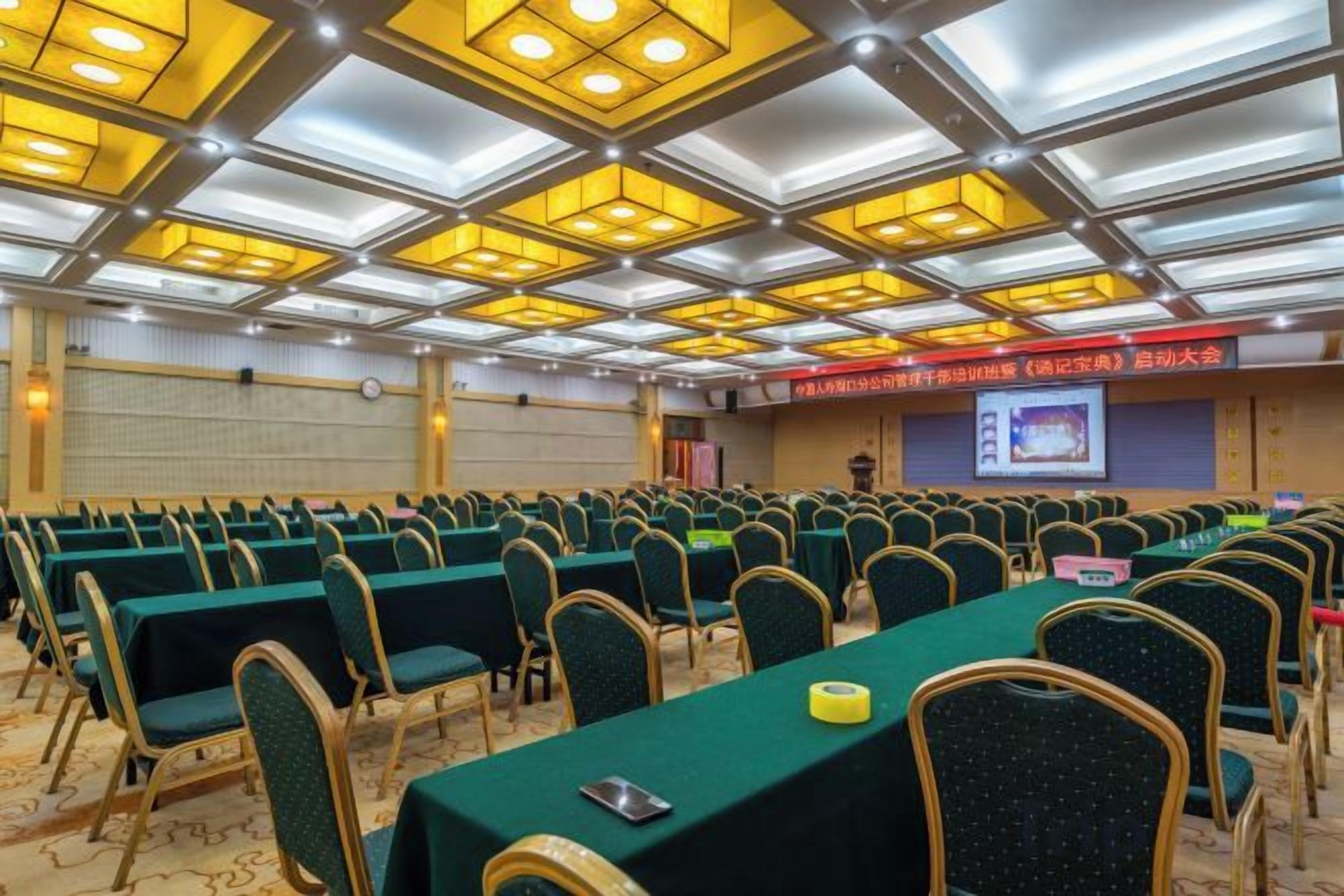 Konferenssisali, jossa on riveissä pöytiä ja tuoleja, esitysnäyttö ja koroke, jossa on puhujapönttö.