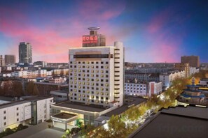 A multi-story hotel with a prominent sign on the roof, surrounded by other buildings and a landscaped area with trees. - Zhoukou Hotel (Zhoukou Wanshunda Department Store) (Zhoukou)