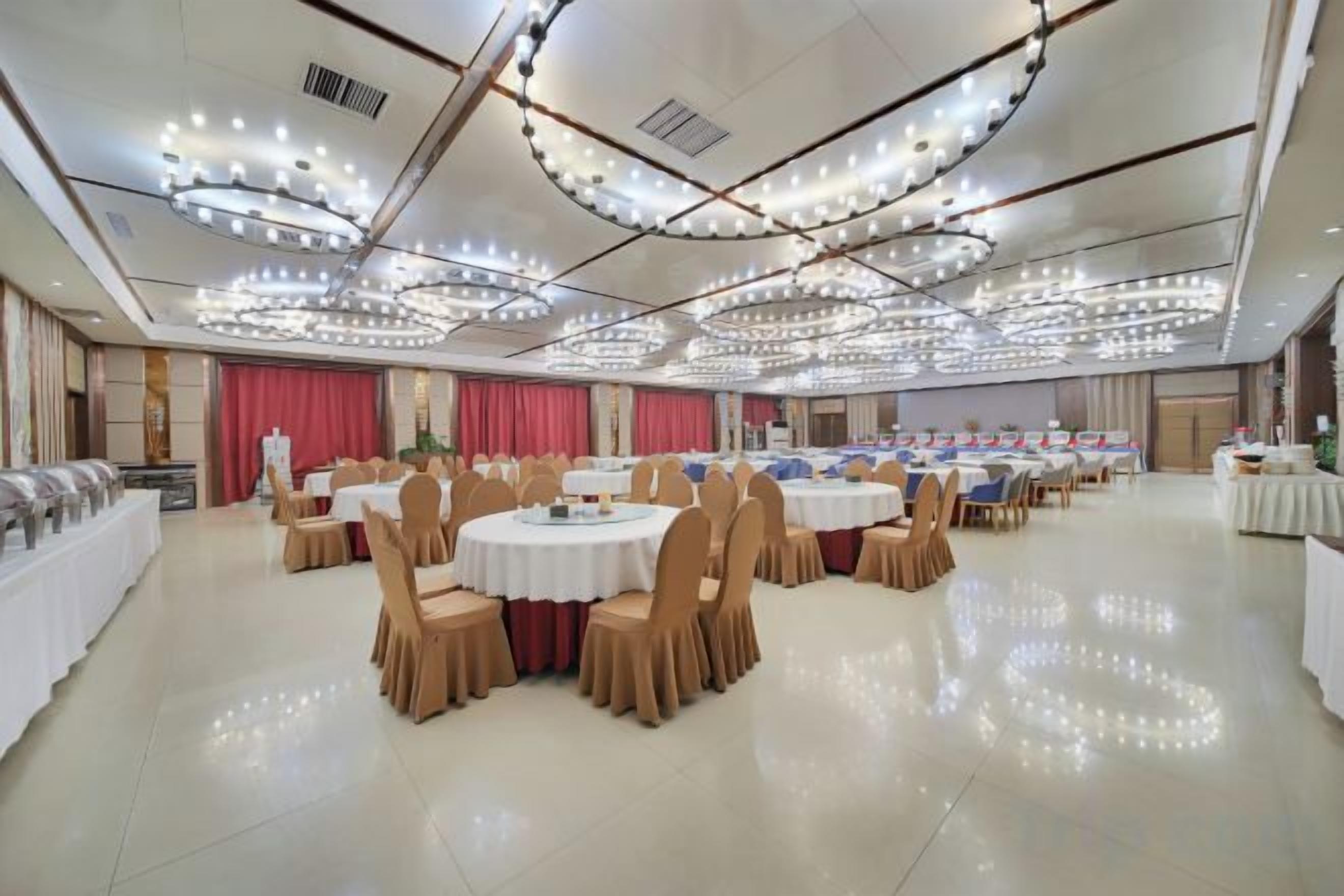 A banquet hall with round tables and chairs, a stage with curtains, and a ceiling with circular light fixtures.
