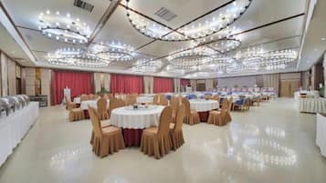 A banquet hall with round tables and chairs, a stage with curtains, and a ceiling with circular light fixtures.