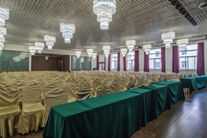 Salão de banquetes com fileiras de cadeiras revestidas em bege, uma longa mesa com toalha verde e vários lustres.