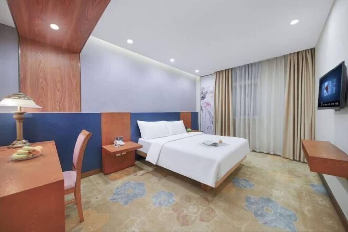 A hotel room with a bed, a desk, a chair, a nightstand, a TV, and a window with curtains.