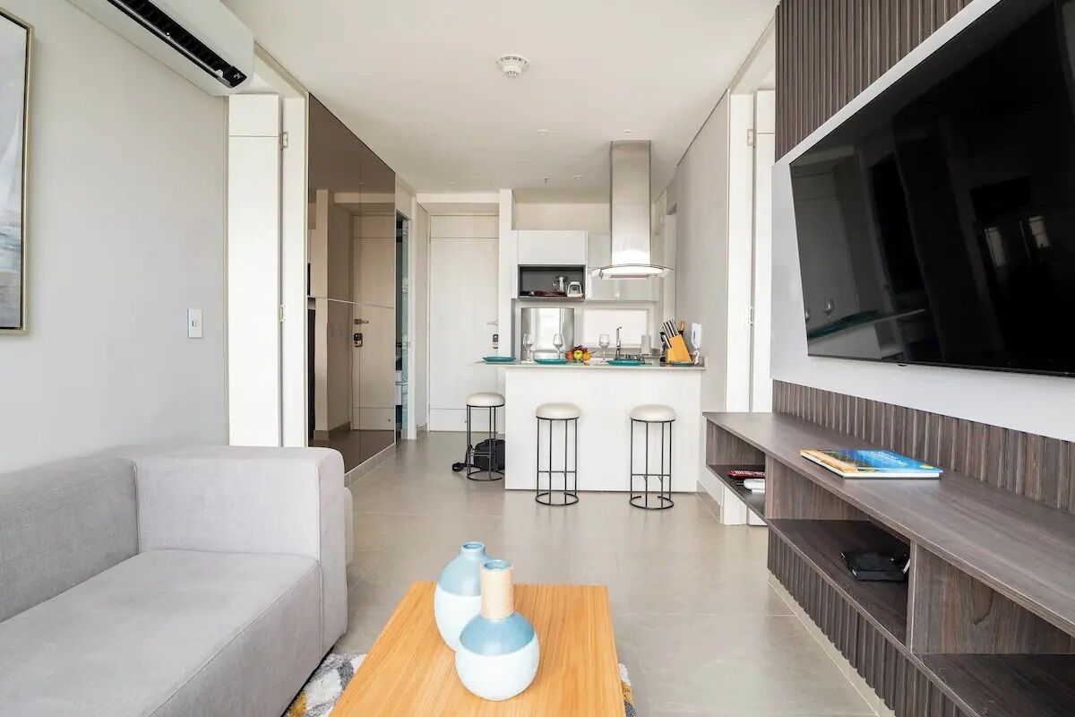 A modern living room with a sofa, a coffee table with vases, a TV on a wooden stand, and a kitchen in the background.