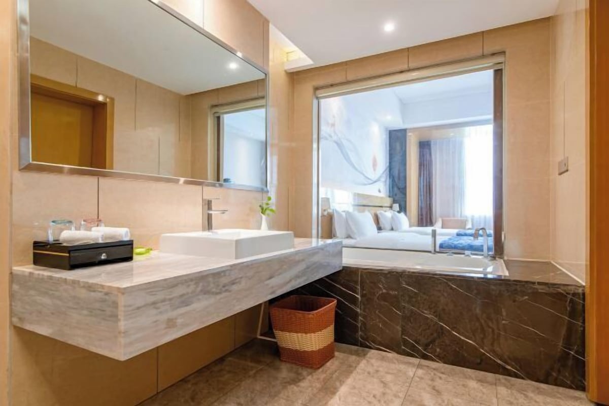A modern bathroom with dual sinks, a large mirror, and a view of a bedroom.
